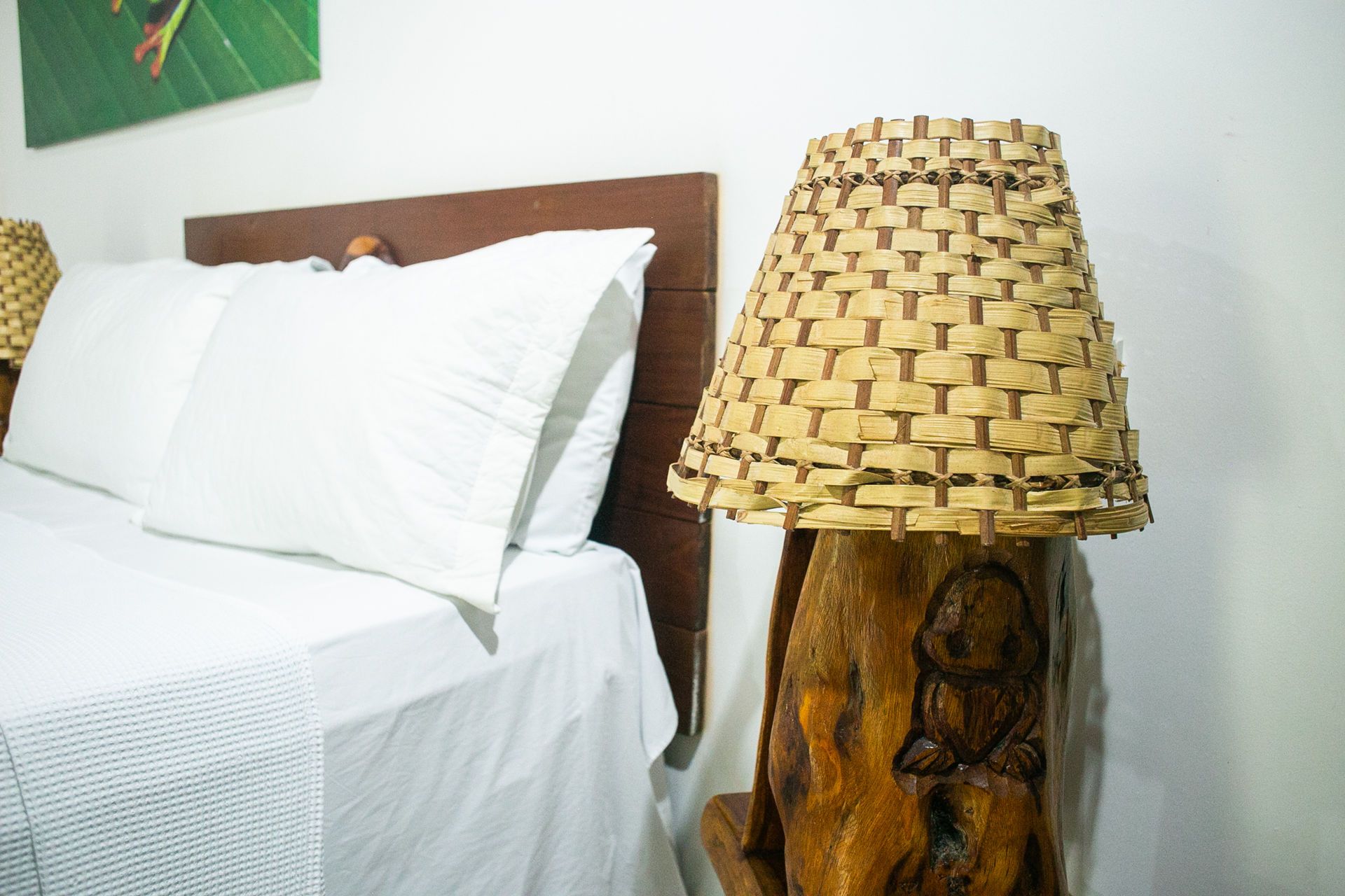 Bedroom scene: bed with white linens, wooden headboard, and a wood-carved lamp with a woven lampshade.