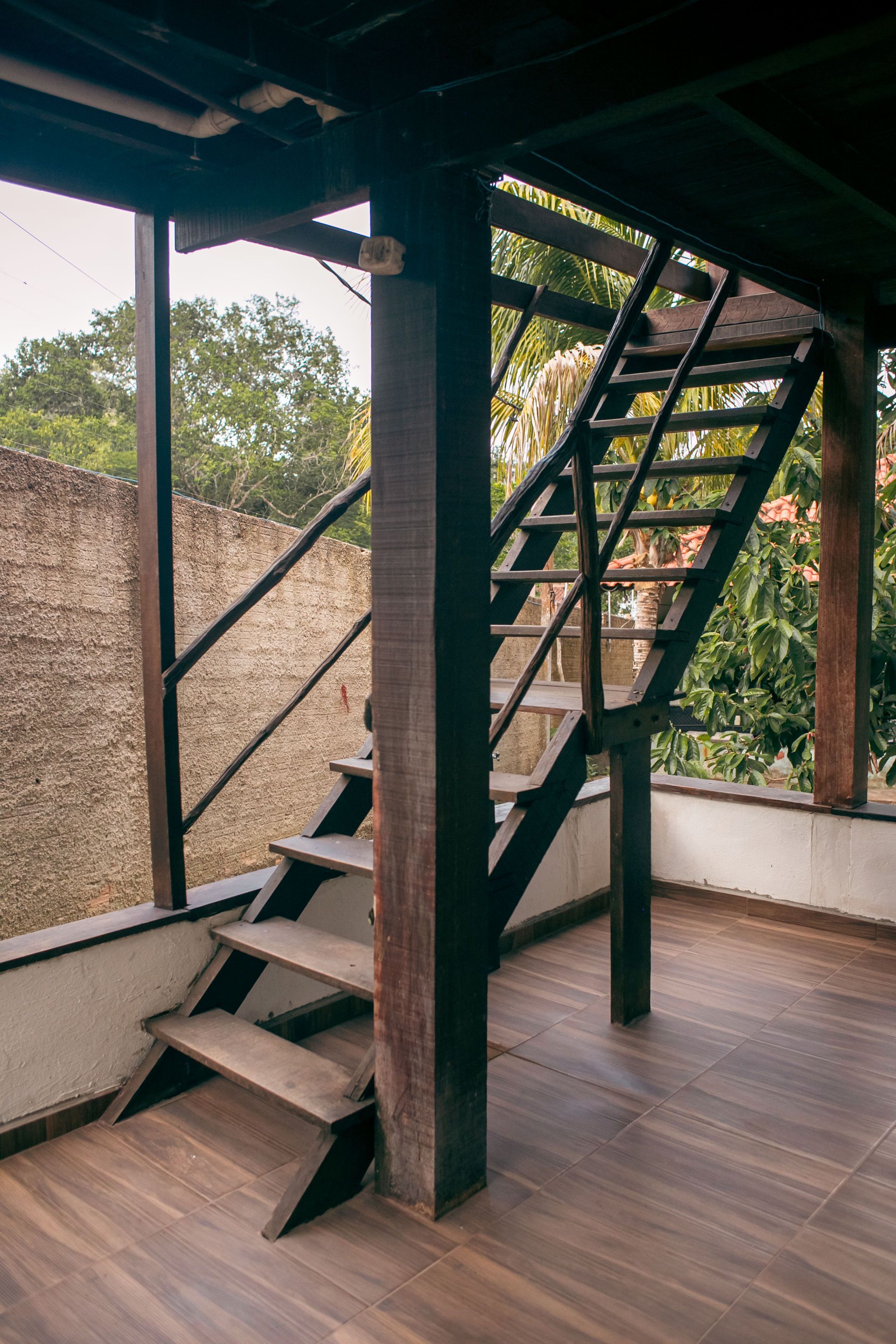 Wooden staircase leading up, set against a brick wall and trees. Brown wood, natural light.
