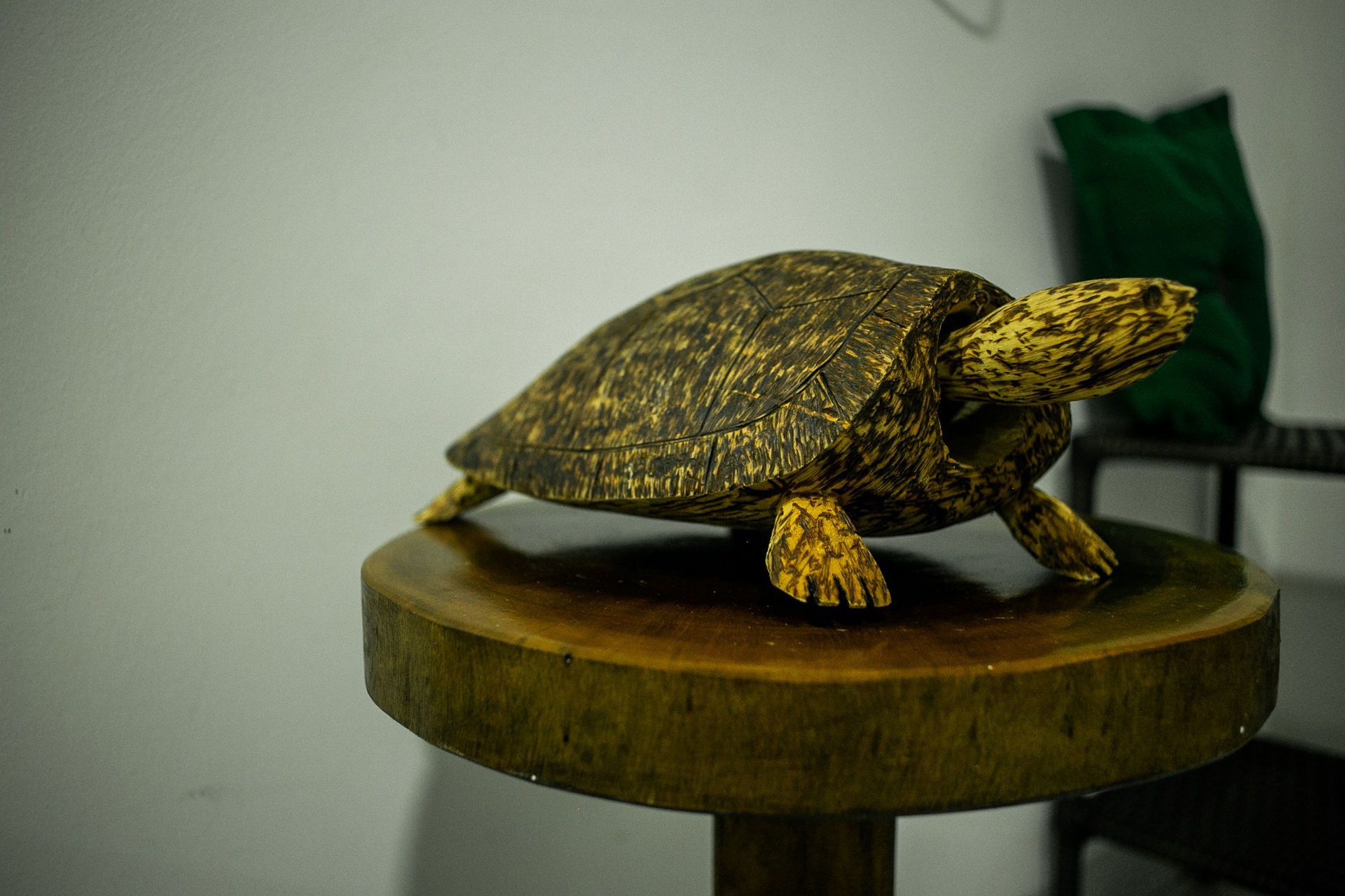 Wooden turtle statue on a round, wooden table, with a green bag and chair in the background.