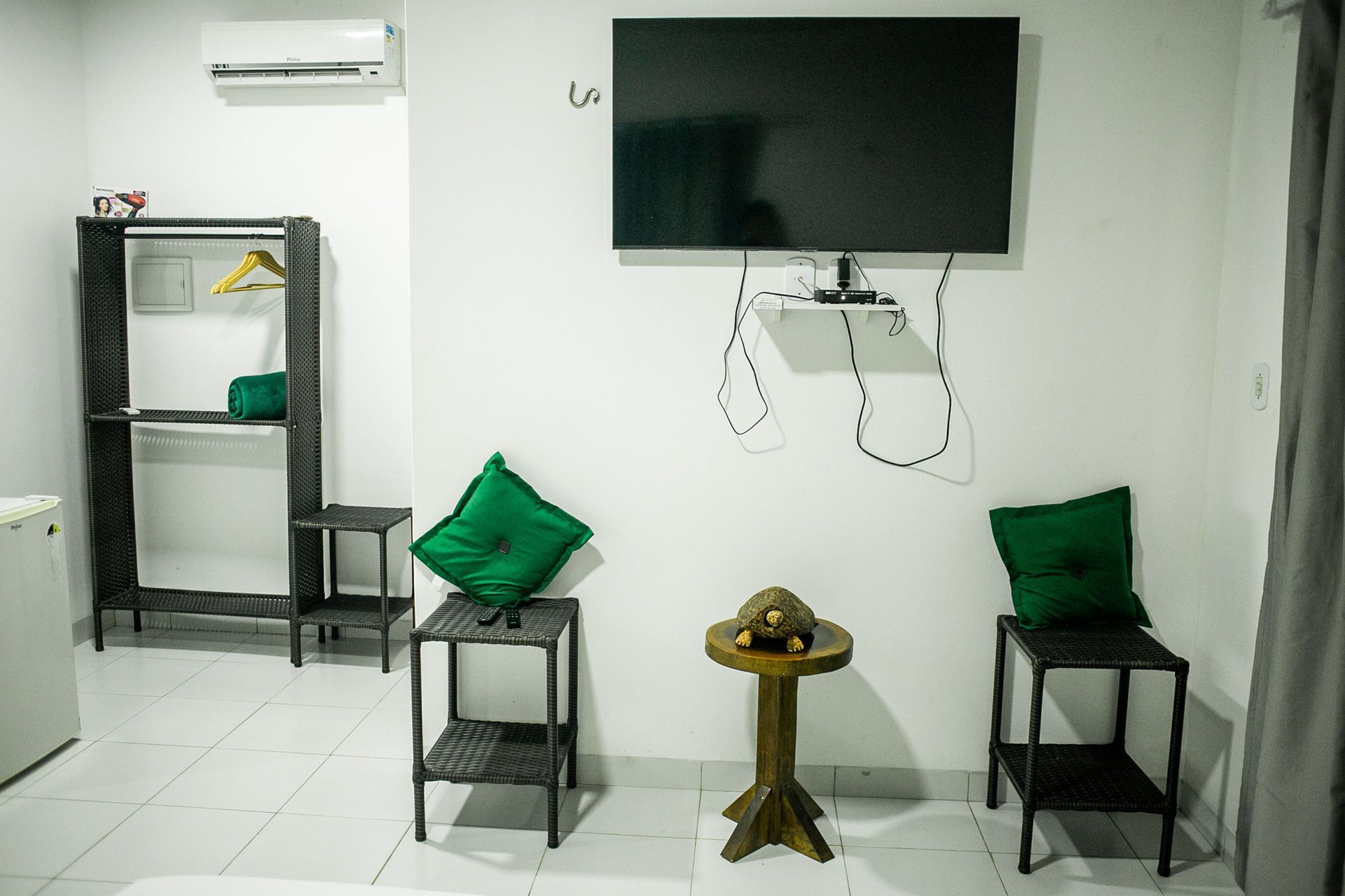 A white-walled room with a TV, open wardrobe, two side tables with green pillows, and an air conditioner.