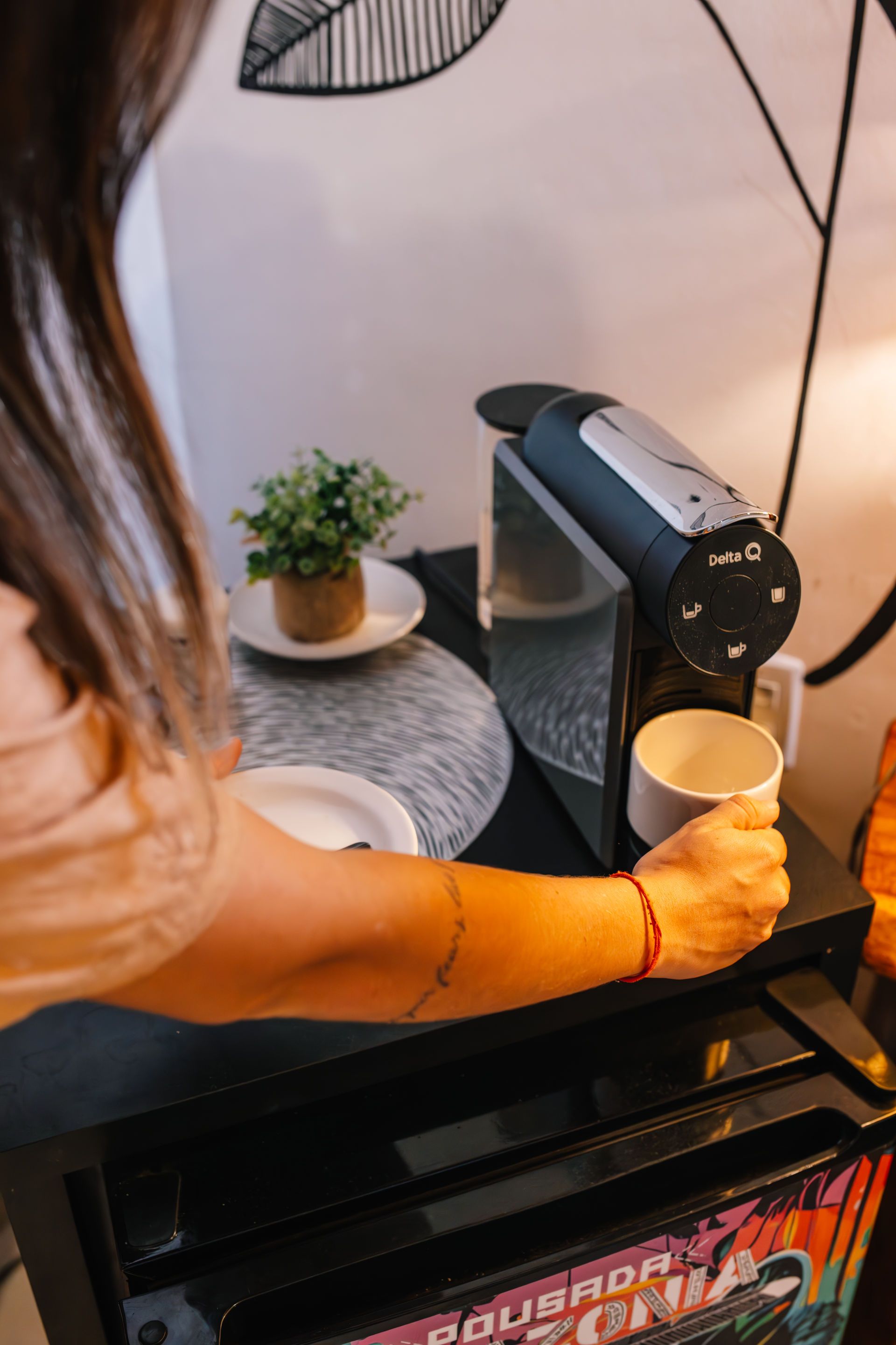 Woman using coffee maker. She holds a mug, next to a plant on a table with decorative art in a room.