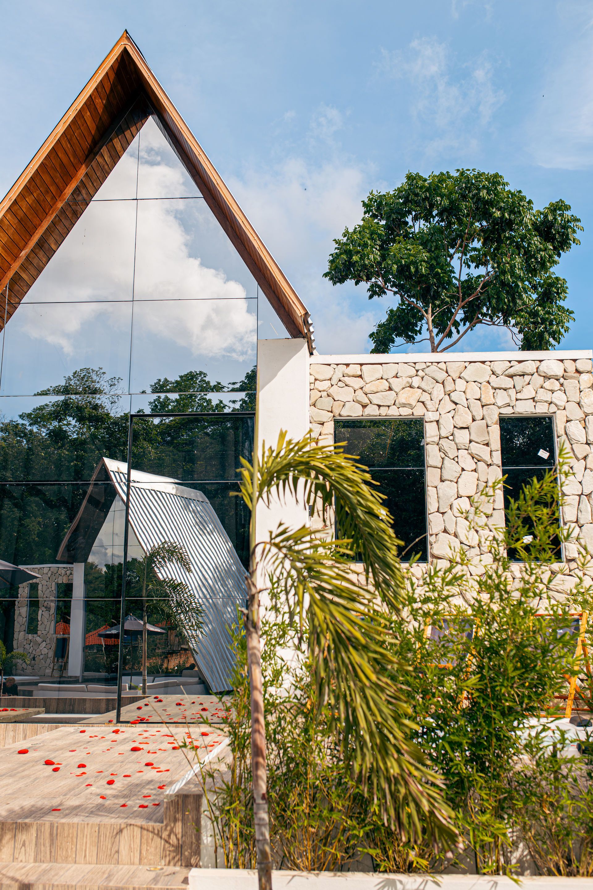 Mirrored-glass triangular building reflects sky and trees, with a stone-textured wall and tropical vegetation.