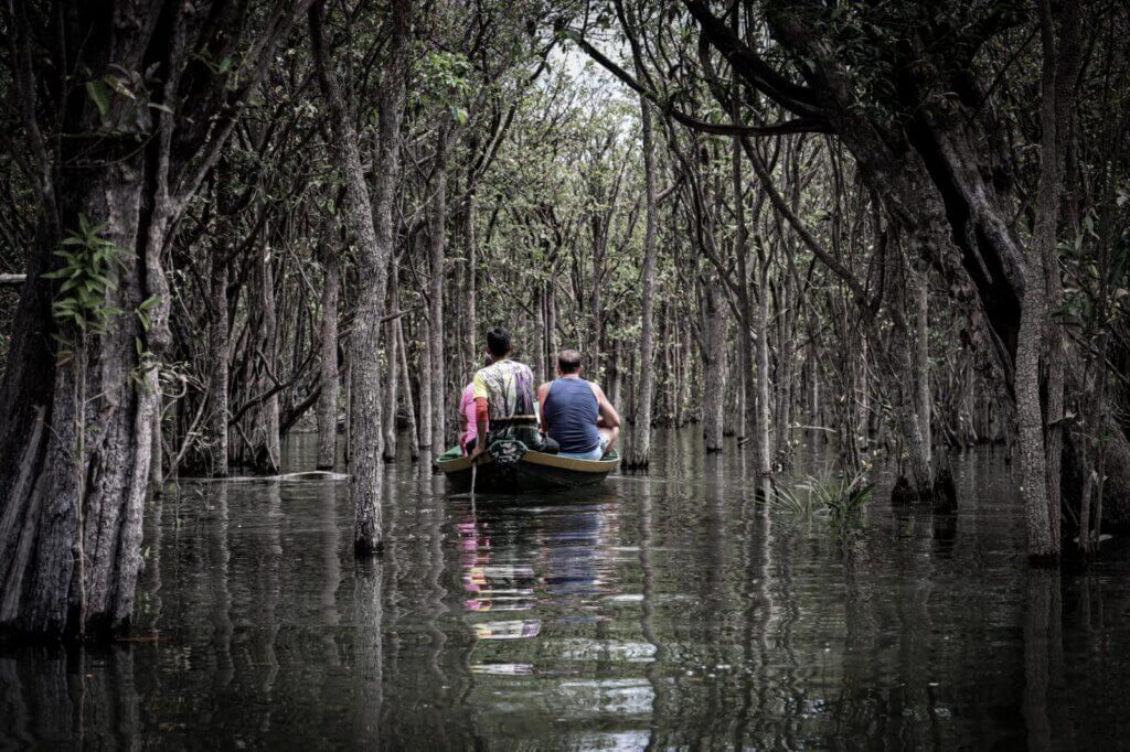 Two people in a boat navigate a dark flooded forest, trees reflected in the still water.
