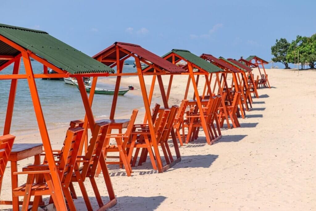 Beachside tables and chairs with orange frames and green/red roofs on a sandy beach.