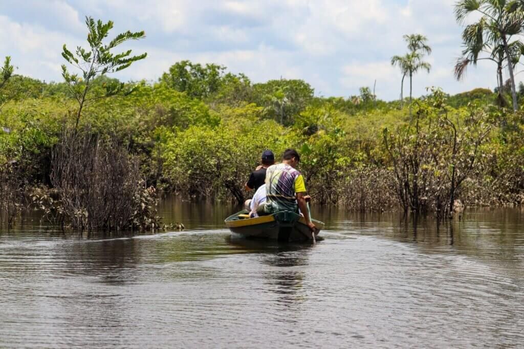 Two people in a small boat navigate a narrow waterway, surrounded by dense green vegetation under a cloudy sky.