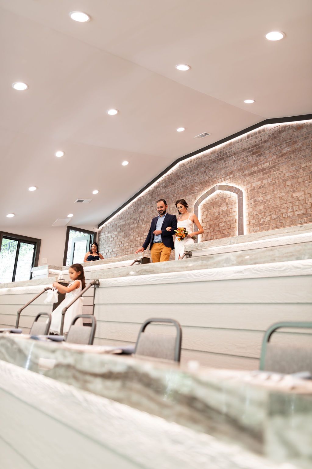 Couple walks down a staircase aisle in a modern, light-filled venue.  A child sits nearby.