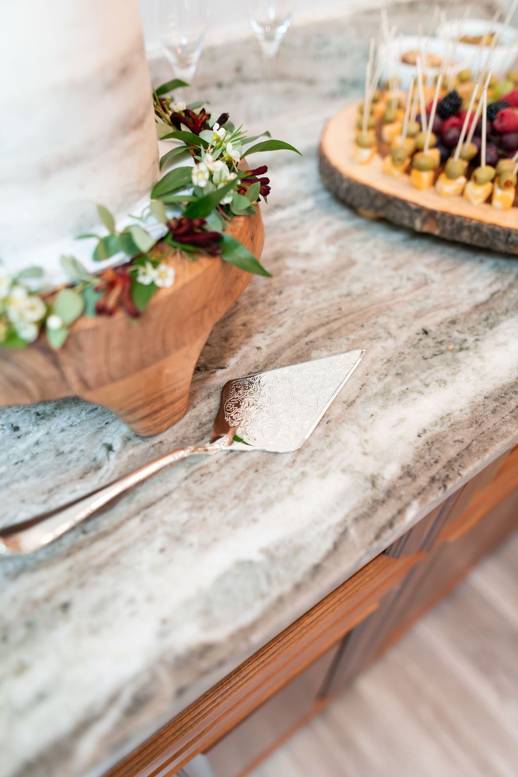 Silver cake server next to a wedding cake on a wooden stand, with appetizers on a wooden board.