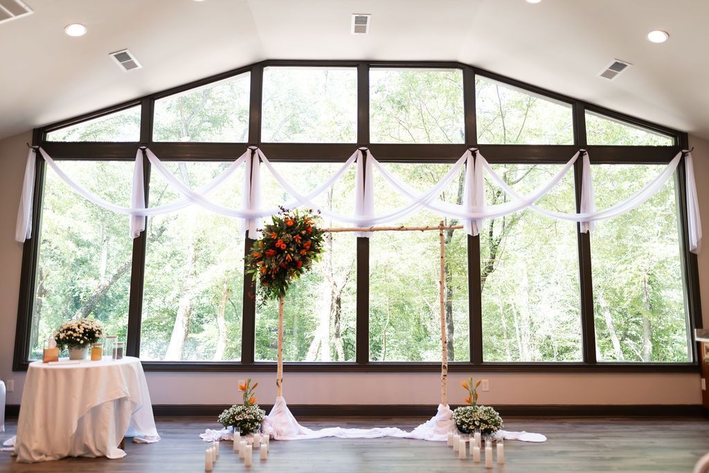 Wedding ceremony setup with arch, flowers, and draped fabric in front of large windows overlooking greenery.