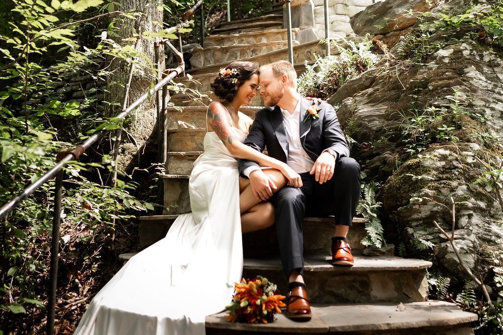 Bride and groom sit on stone steps, embracing; surrounded by greenery.