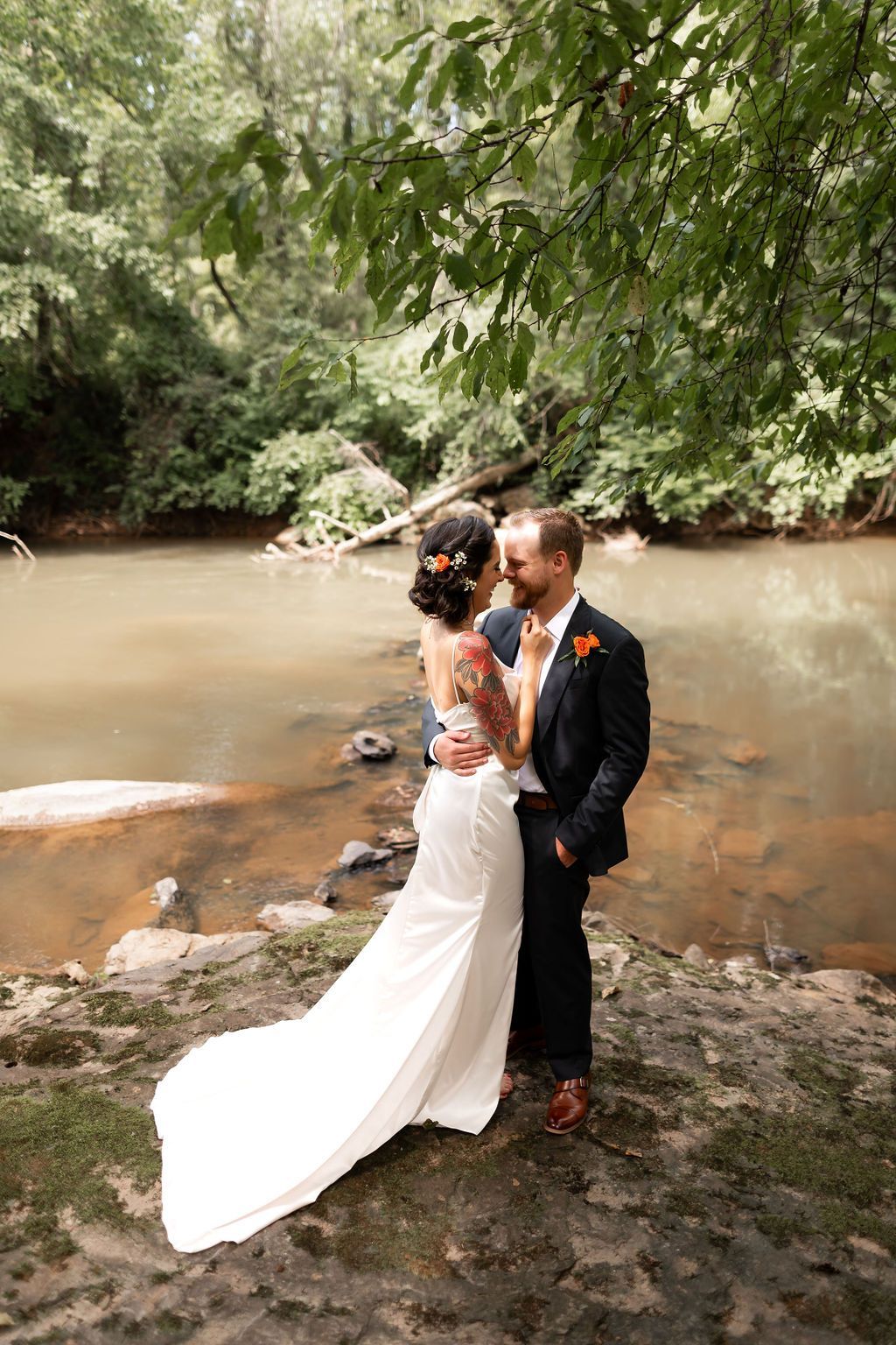 Bride and groom embrace by a river in a forest, bride in a white dress, groom in a navy suit.
