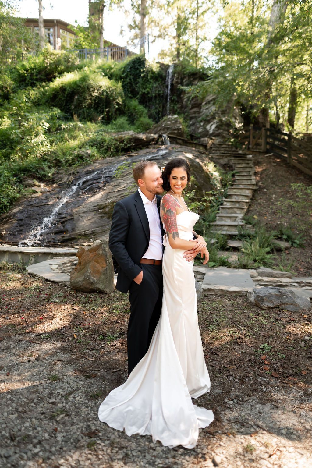 Bride and groom embracing outdoors by a small waterfall; bride in white dress with arm tattoos, groom in a suit.