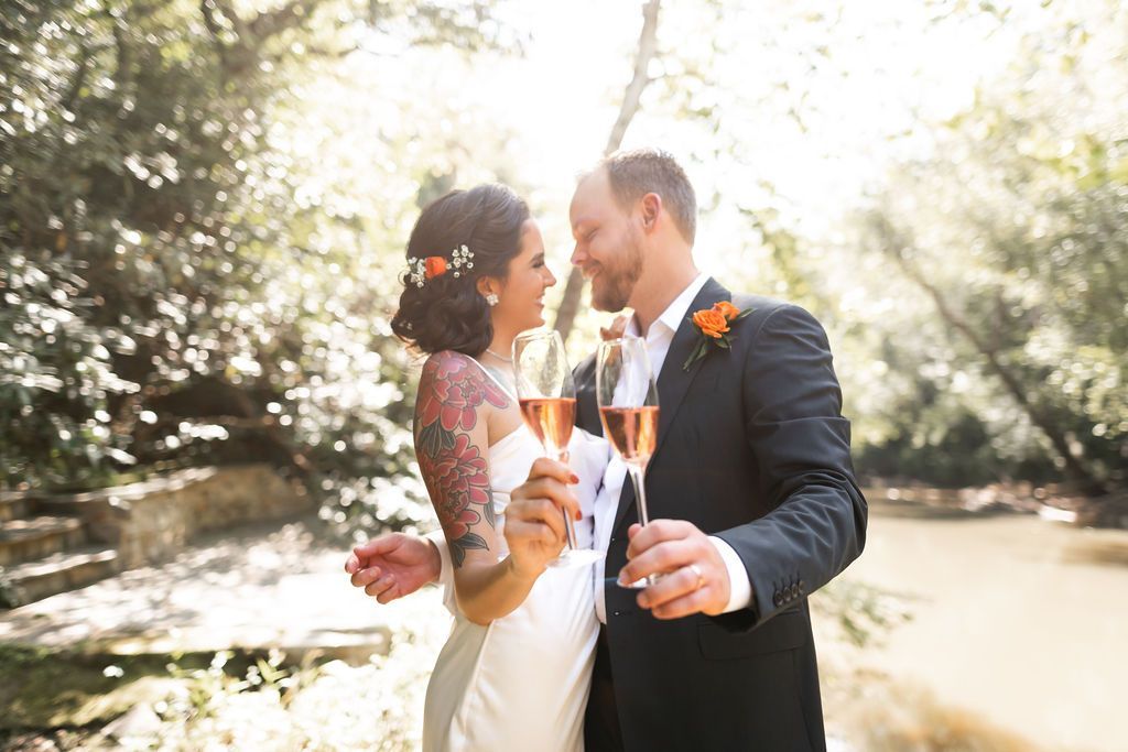Bride and groom toast with champagne, smiling, near a river, in a sunny outdoor setting.