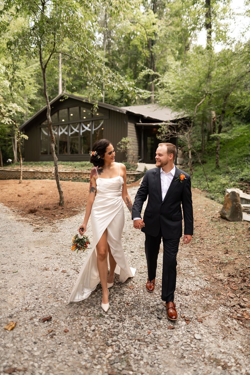 Bride and groom walk hand-in-hand, smiling, toward cabin. She wears white dress, he wears navy suit. Forest setting.