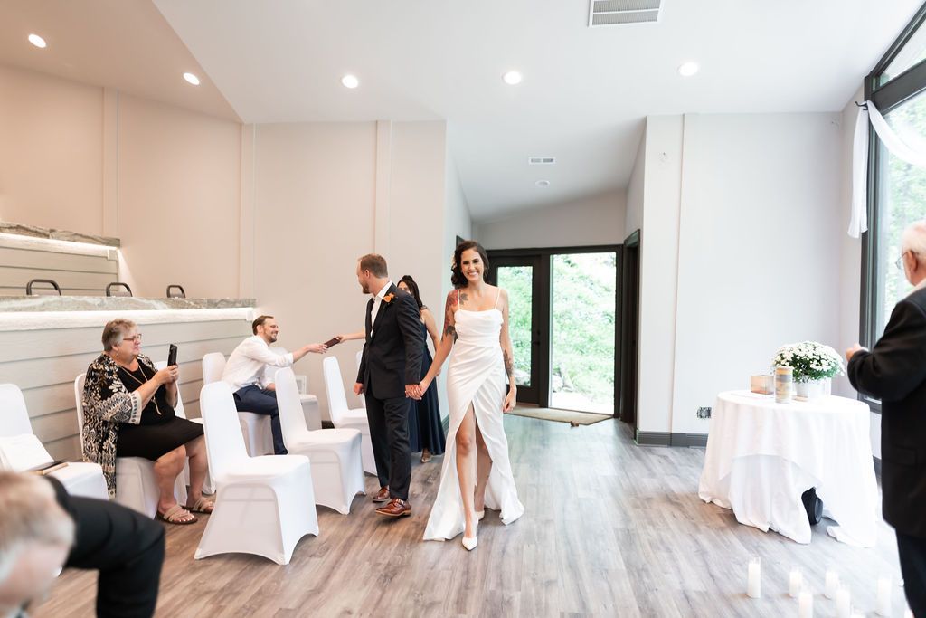 Bride and groom walking hand-in-hand after a ceremony. A few guests, and a table with flowers are also present.