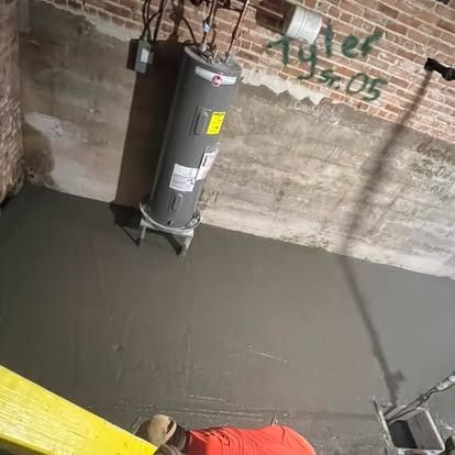 A man is laying concrete in a basement next to a water heater.