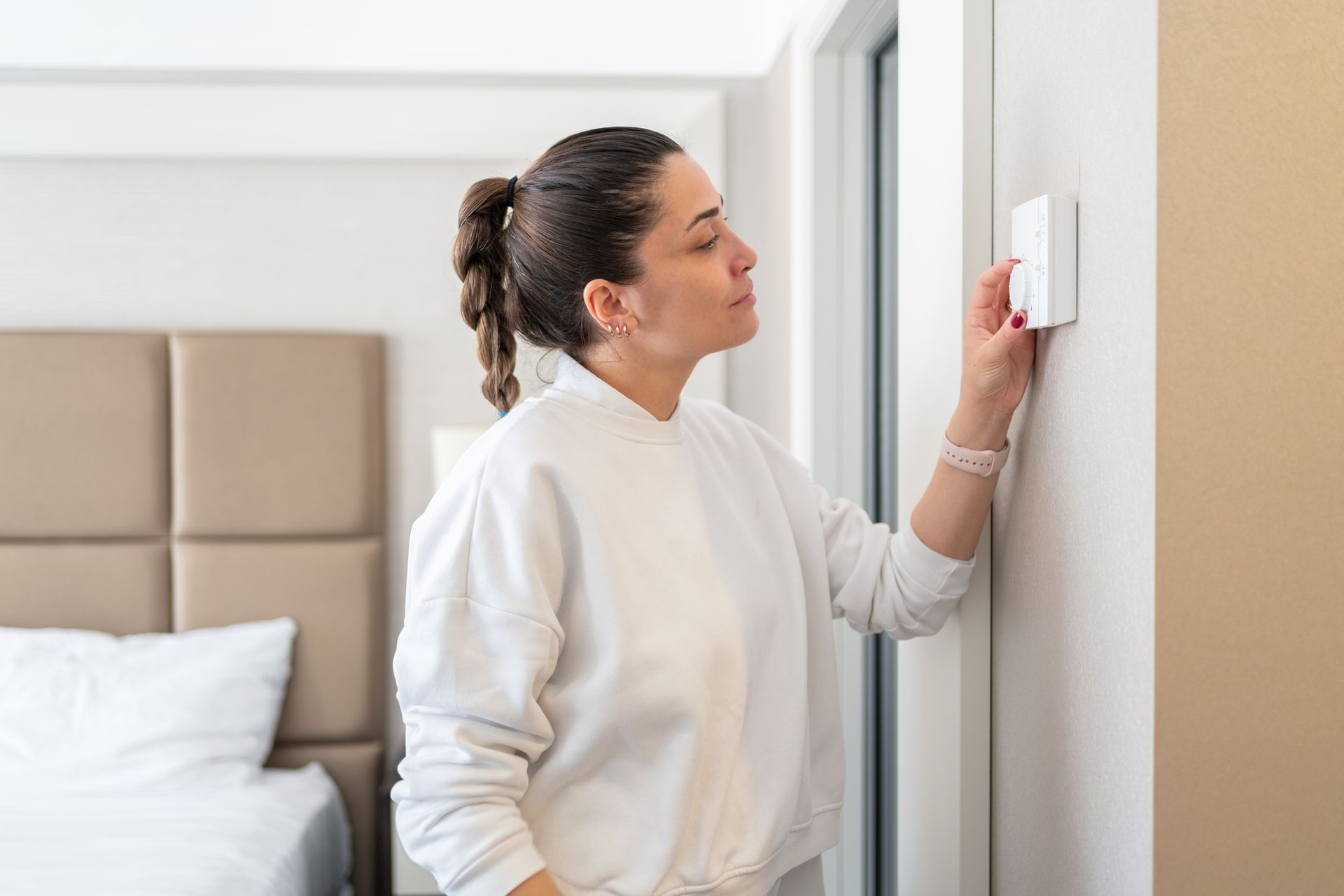 Una donna regola un termostato a parete in una camera da letto con letto e pareti bianche.