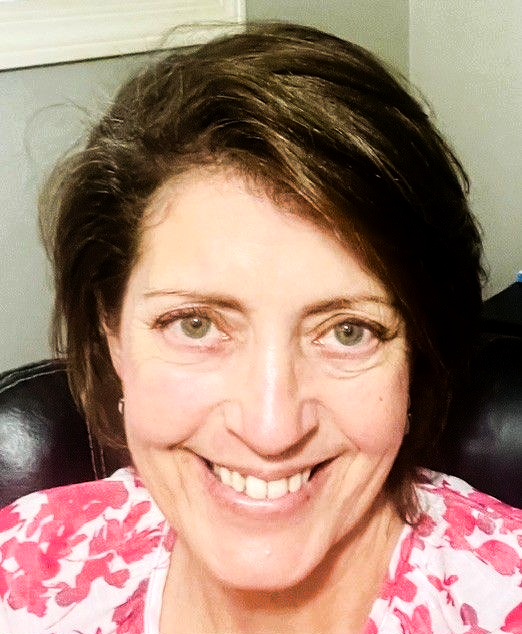 A person with shoulder-length, layered dark hair smiling at the camera, wearing a pink and white floral-print top.