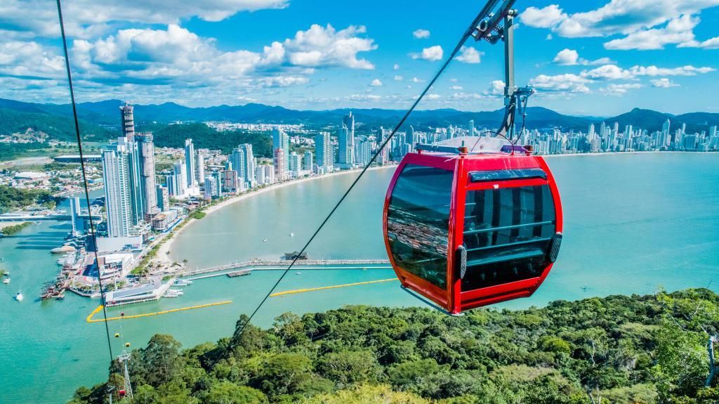 Bondinho sobre a baía de Balneário Camboriú, com skyline de prédios, mar azul e morro verde em dia ensolarado.
