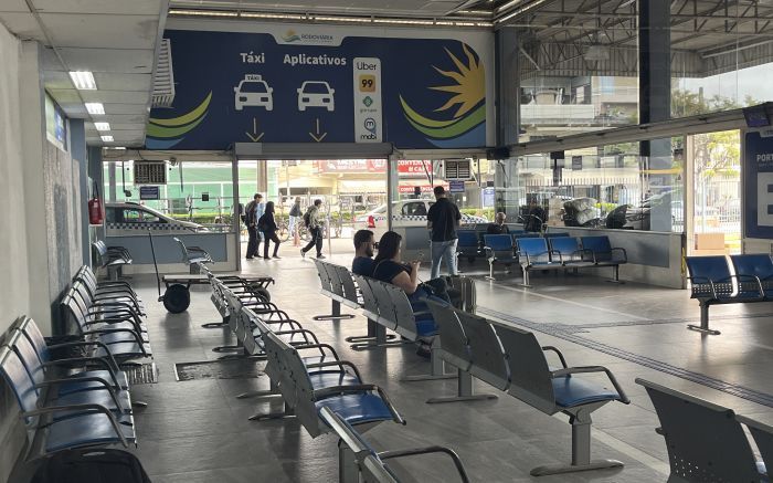 Fileiras de bancos azuis na rodoviária sob uma placa que direciona para táxis e aplicativos de transporte.