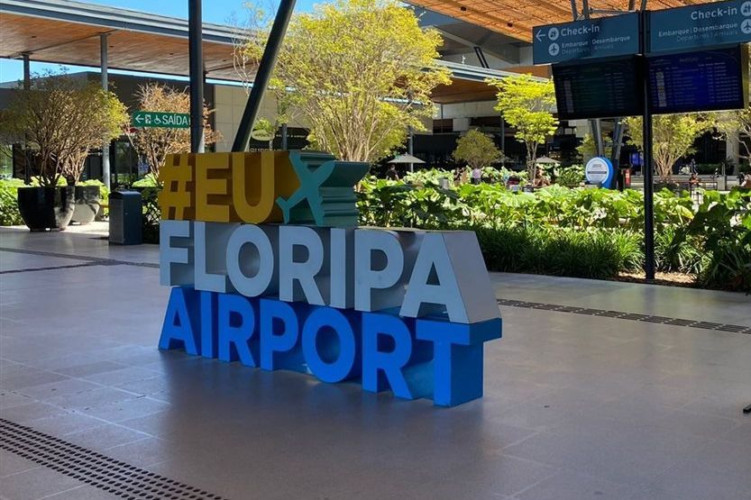 placa colorida “#EUFLORIPAAIRPORT” em aeroporto cercado por jardins e árvores.