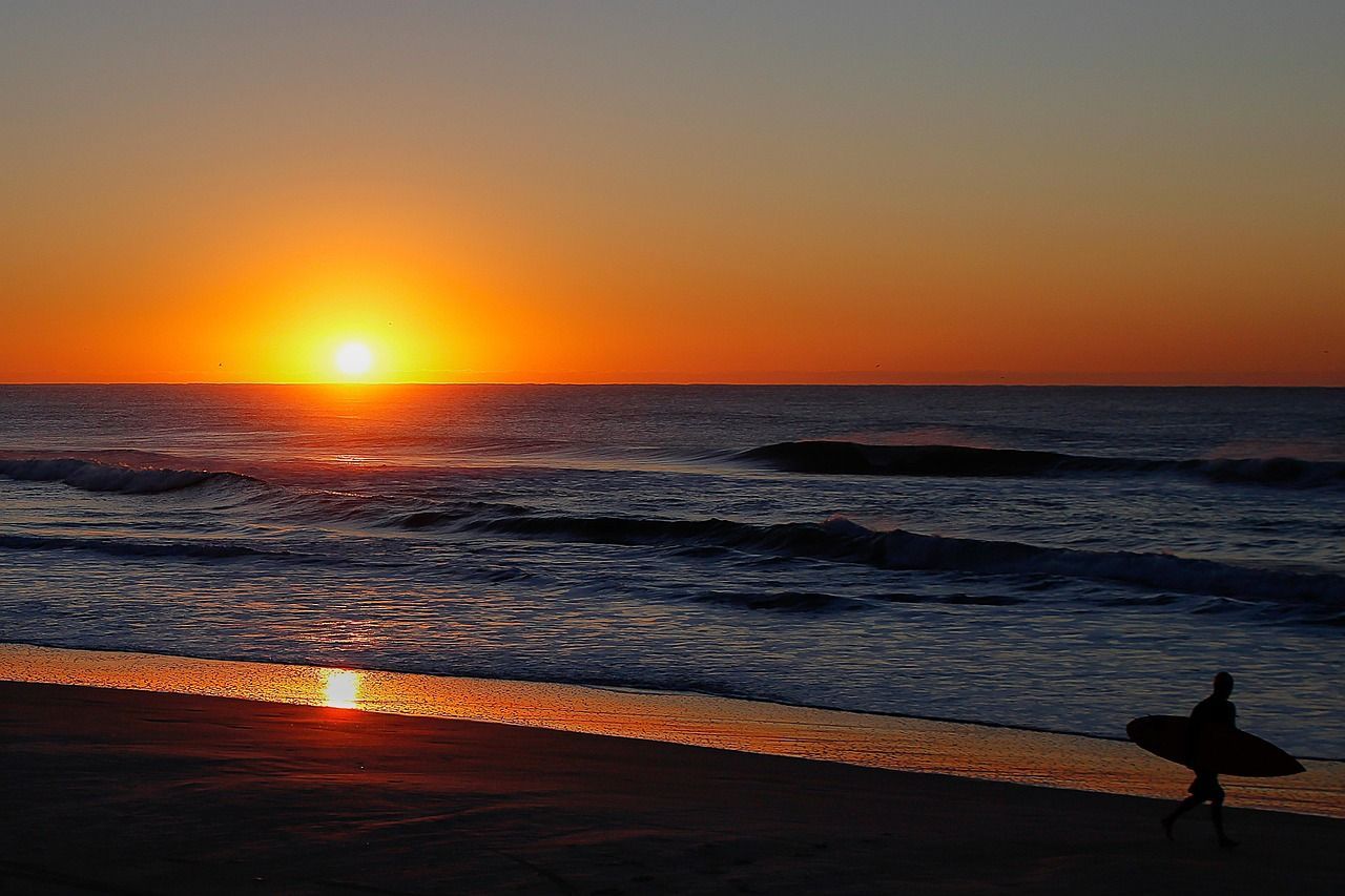 Surfista caminhando na praia ao amanhecer com o sol nascendo no horizonte.