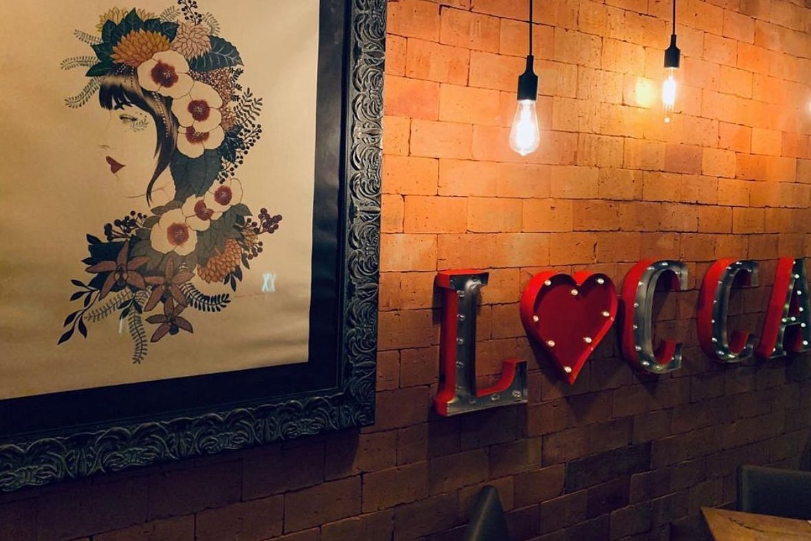 Ambiente com parede de tijolos, quadro artístico de mulher com flores e letreiro iluminado com a palavra LUCCA
