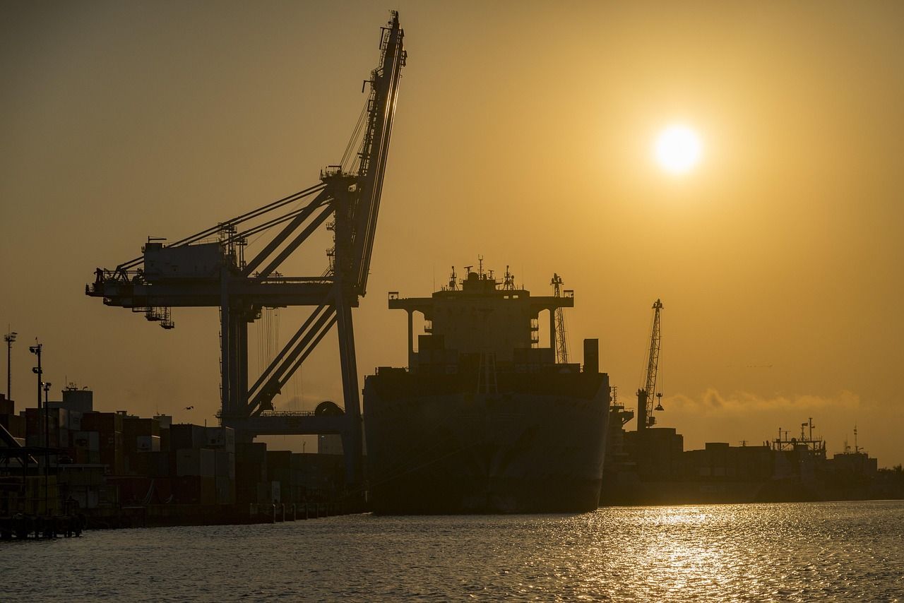 Porto ao entardecer, com navios cargueiros e guindastes silhuetados contra o pôr do sol.