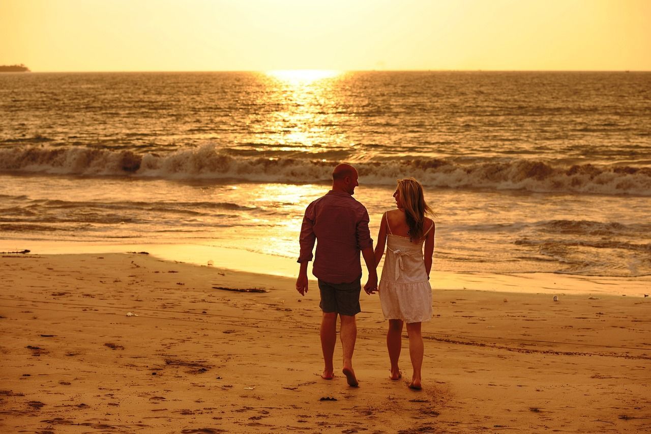 Casal caminha de mãos dadas pela praia ao entardecer.
