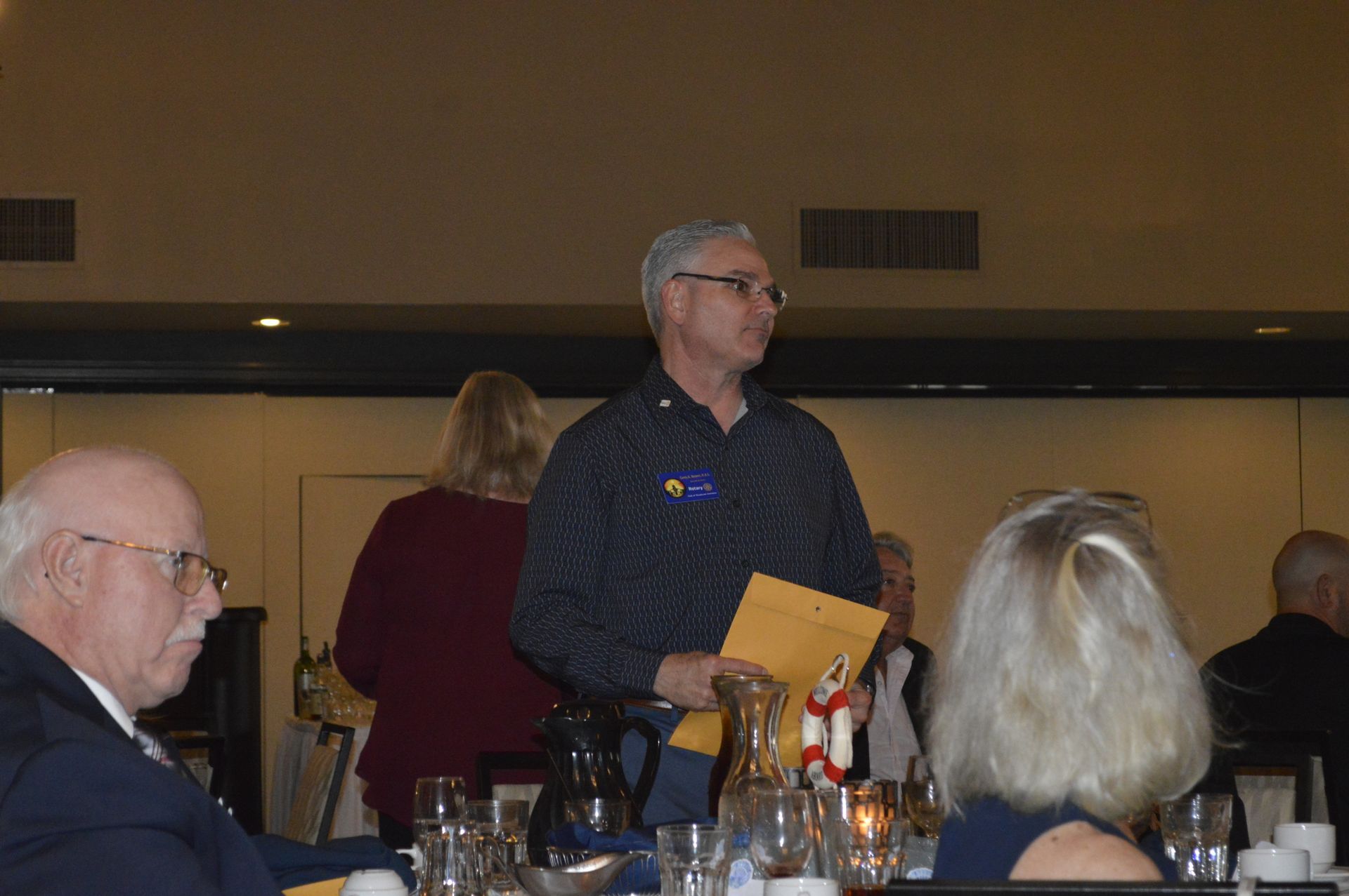 A man is standing in front of a group of people at a table holding a piece of paper.