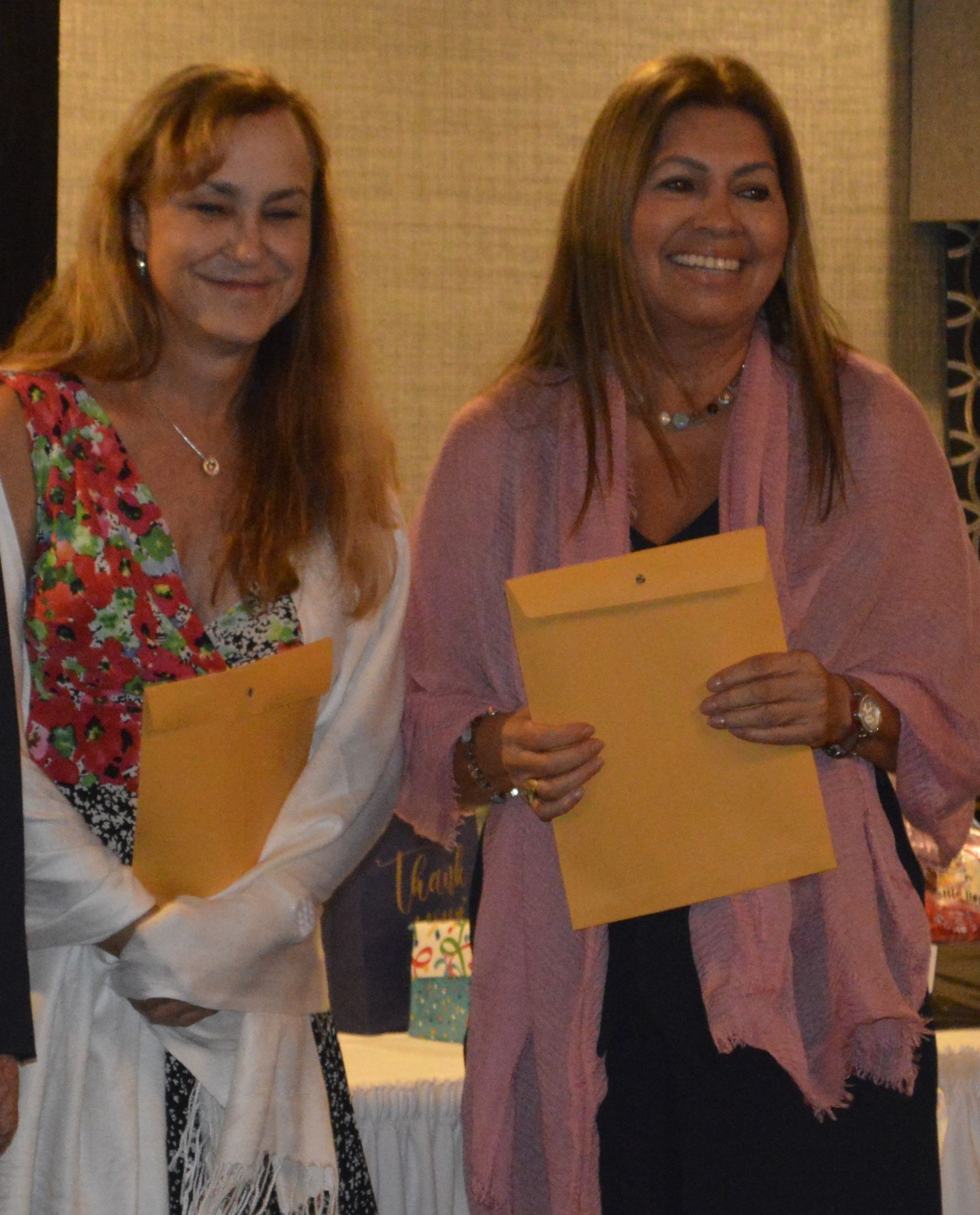 Two women standing next to each other holding envelopes and smiling