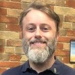 A man with a beard is smiling in front of a brick wall.