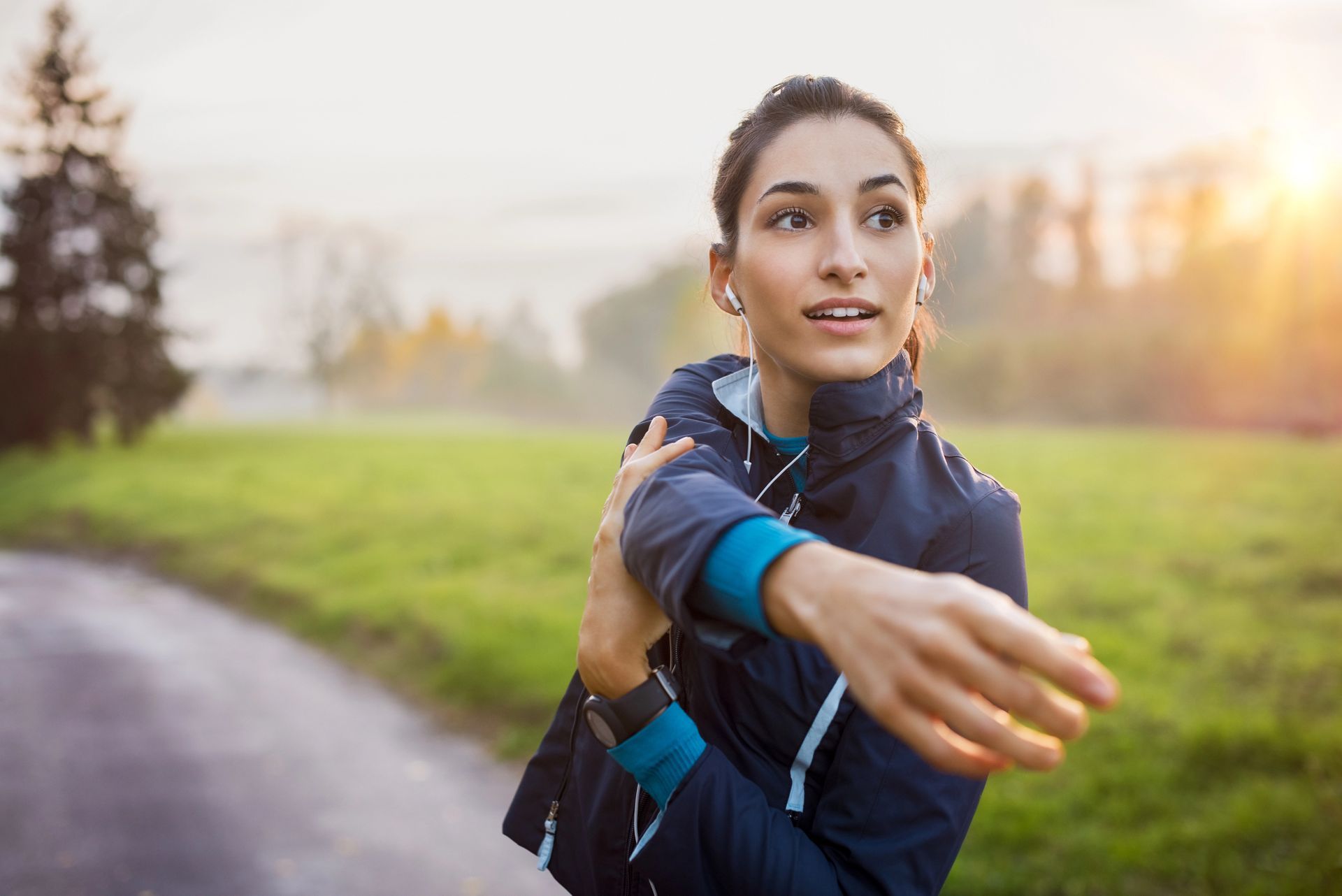 Woman stretching outdoors on a path at sunrise, wearing a dark athletic jacket with blue accents.
