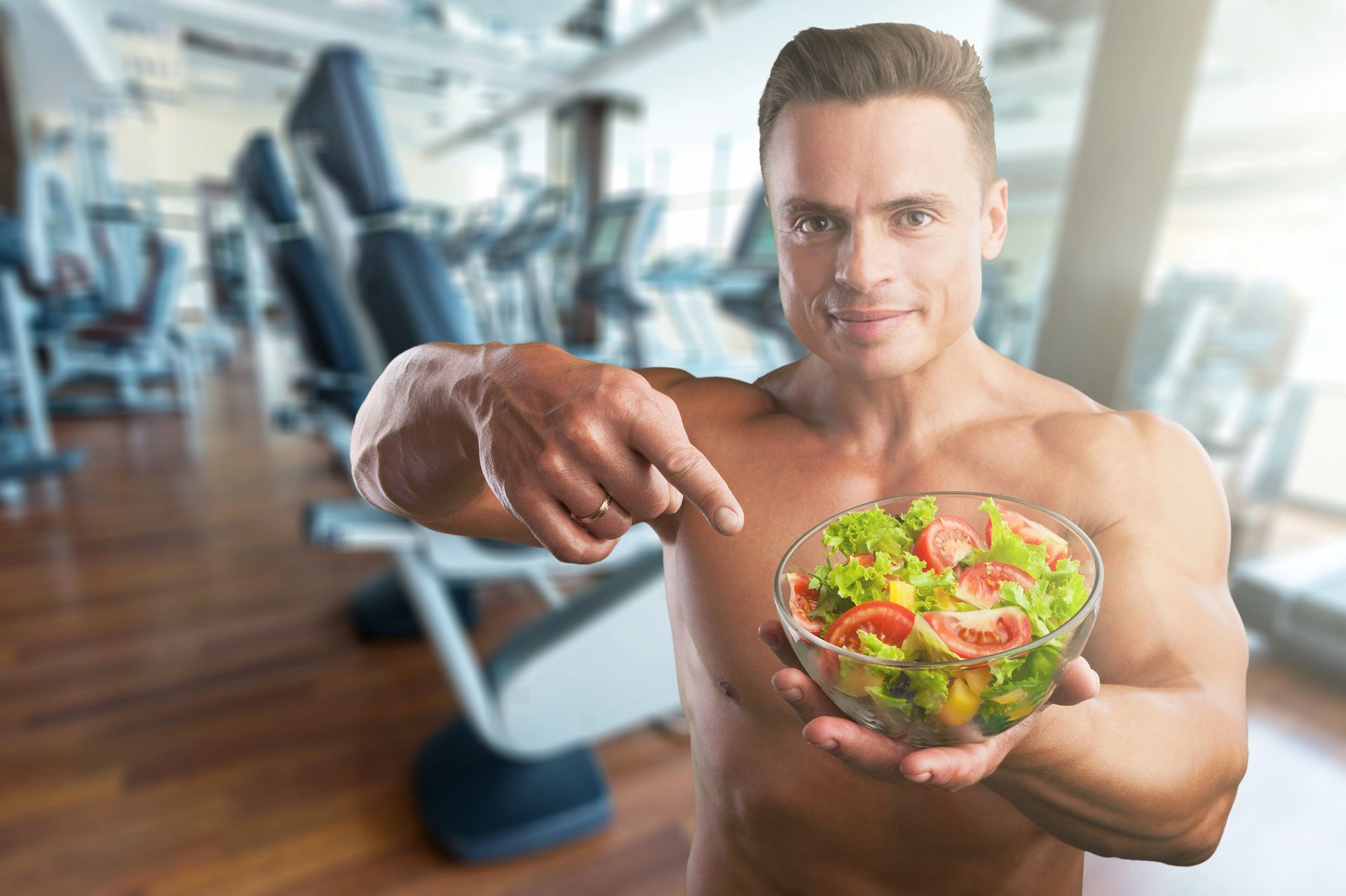 Shirtless man in a gym holding a salad bowl and pointing at it