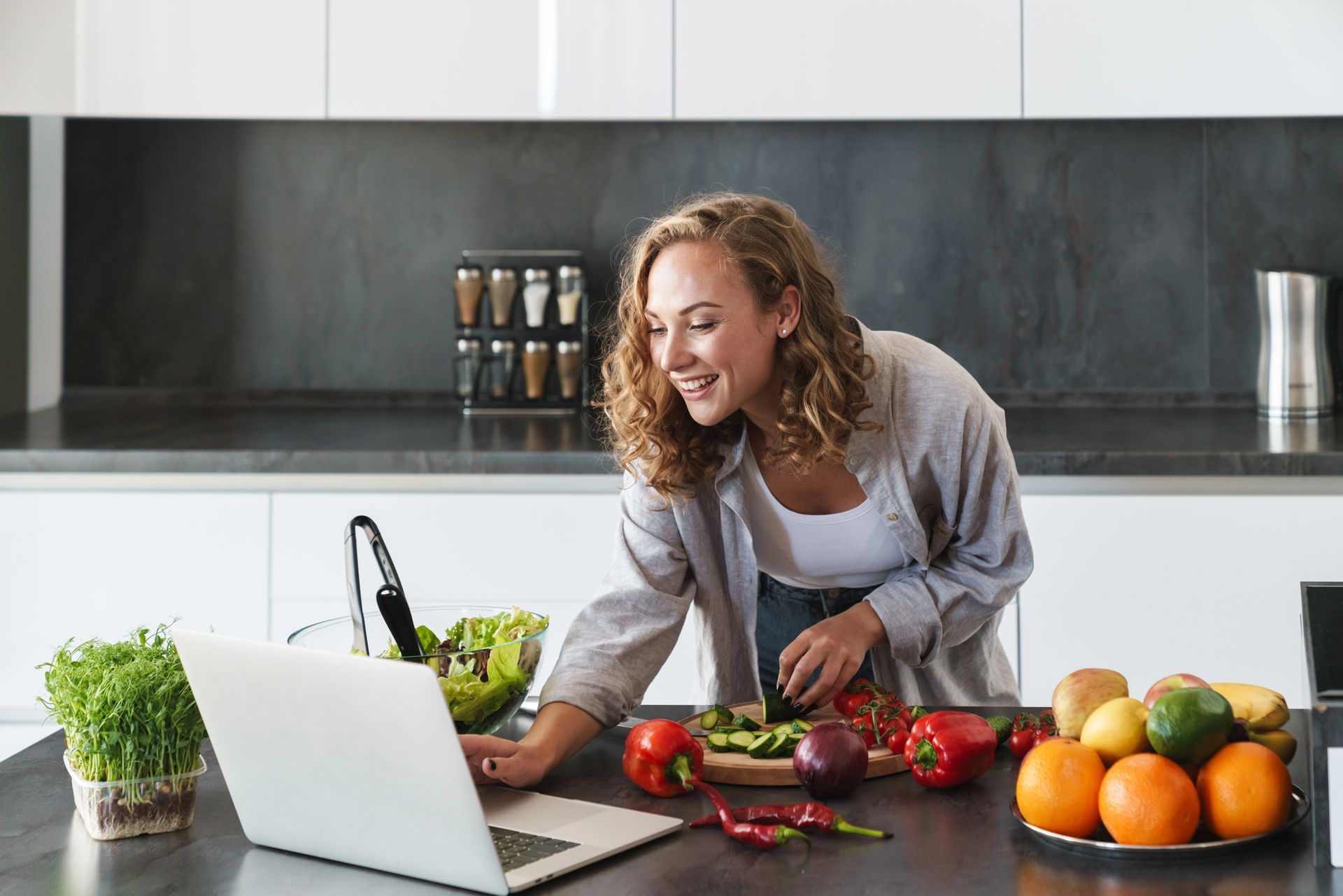 Woman preparing vegetables in a bright kitchen beside a laptop and fruit bowl