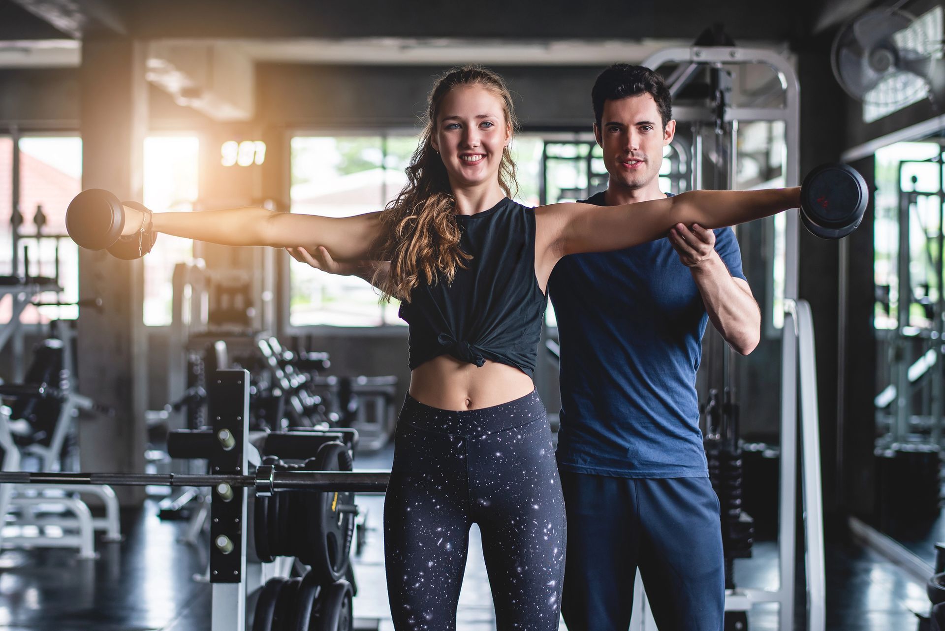 Two people posing with a barbell in a gym, smiling under bright lights.