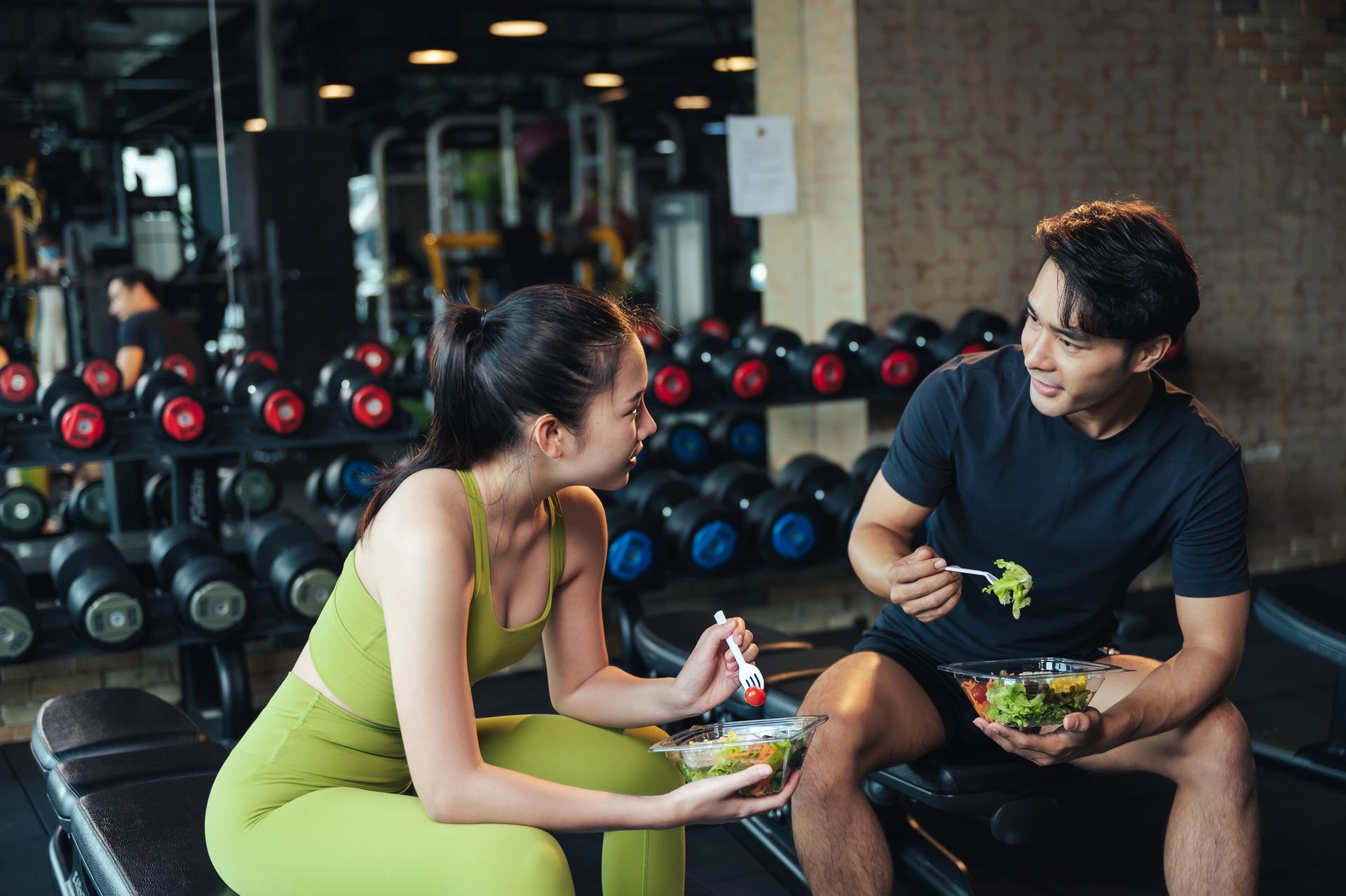 Two people eating salad in a gym, seated near rows of dumbbells.