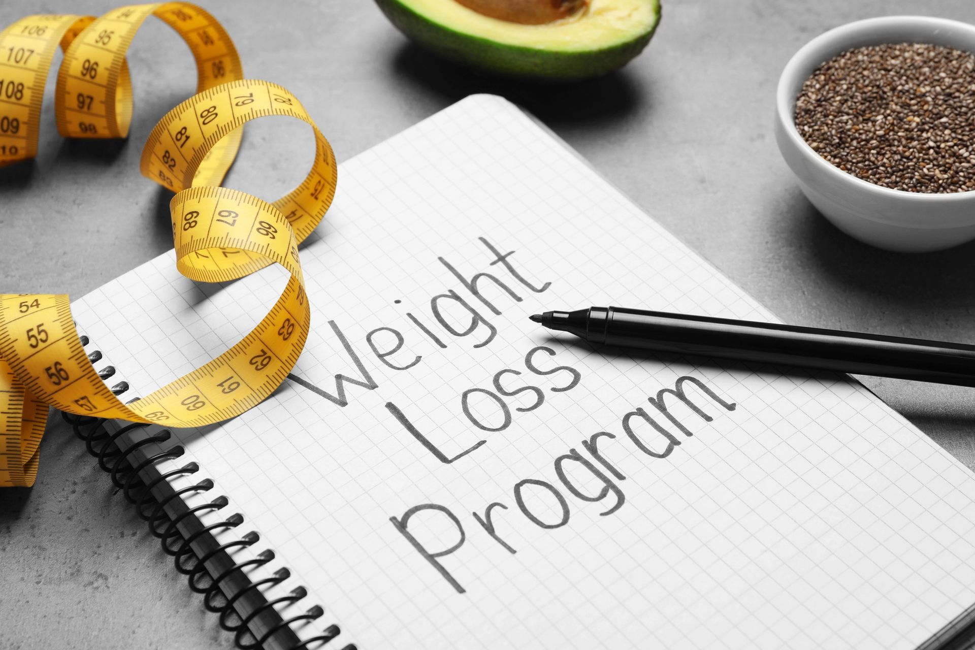 Weight loss program notebook with measuring tape, avocado, chia seeds, and pen on a table