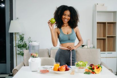 Woman preparing fruit in a bright kitchen, holding a green apple beside a blender and fresh ingredients.