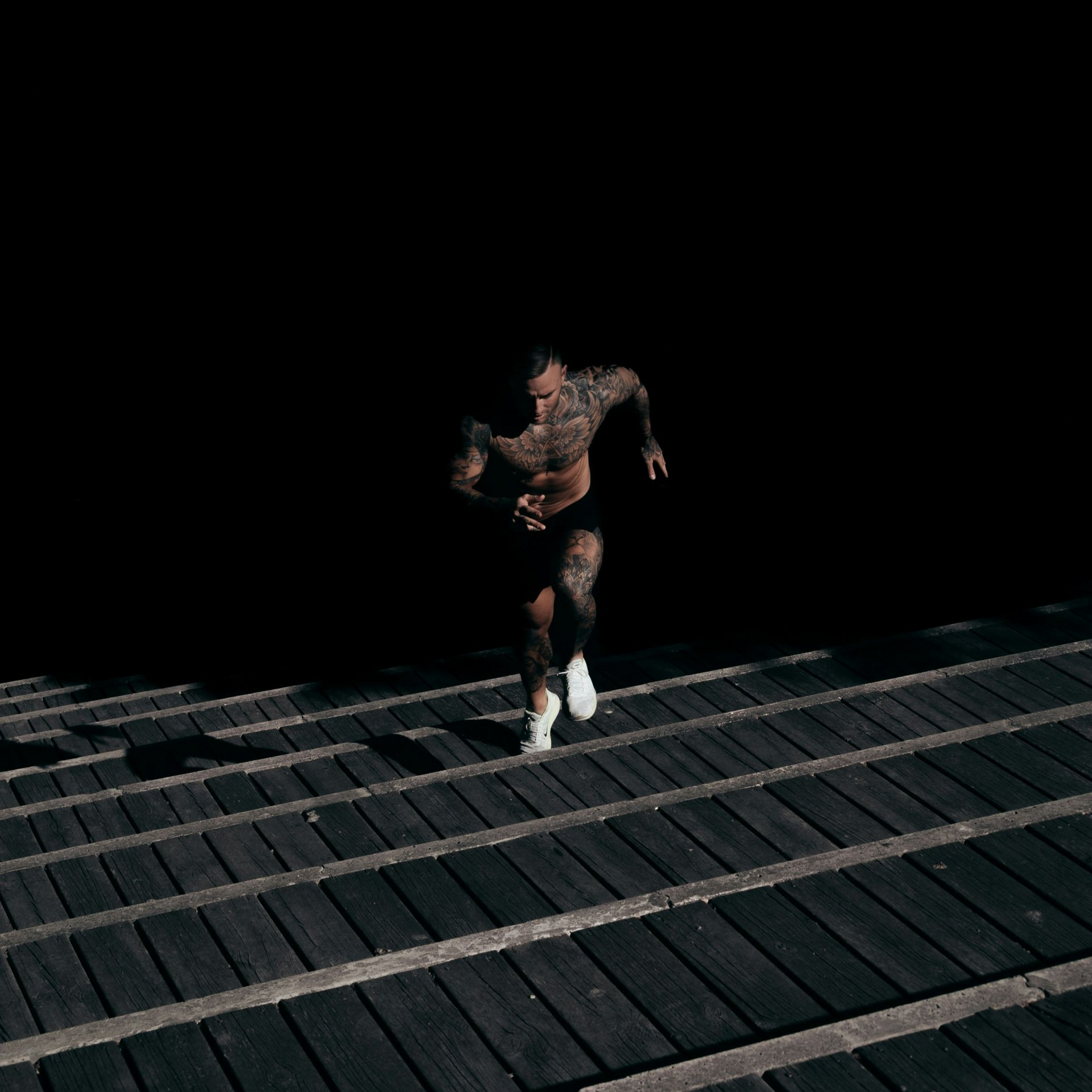 Runner on a wooden boardwalk at night, emerging from darkness with a forward stride.