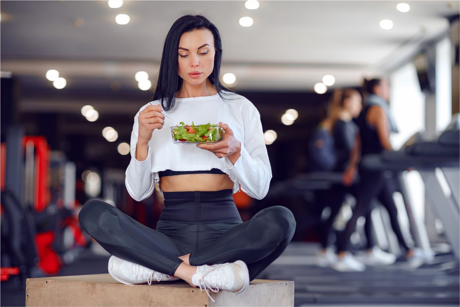 Woman eating salad while seated cross-legged in a gym