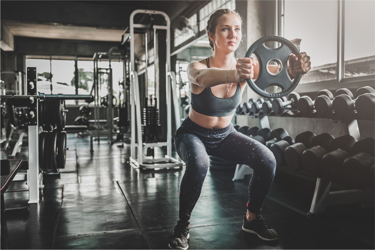 Woman doing a weighted squat in a gym beside dumbbells and exercise equipment