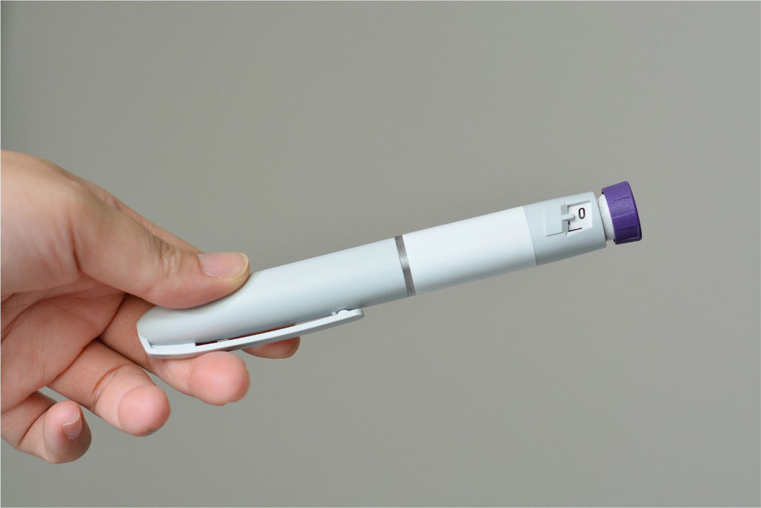 Hand holding a white pen-like medical device with a purple cap against a gray background