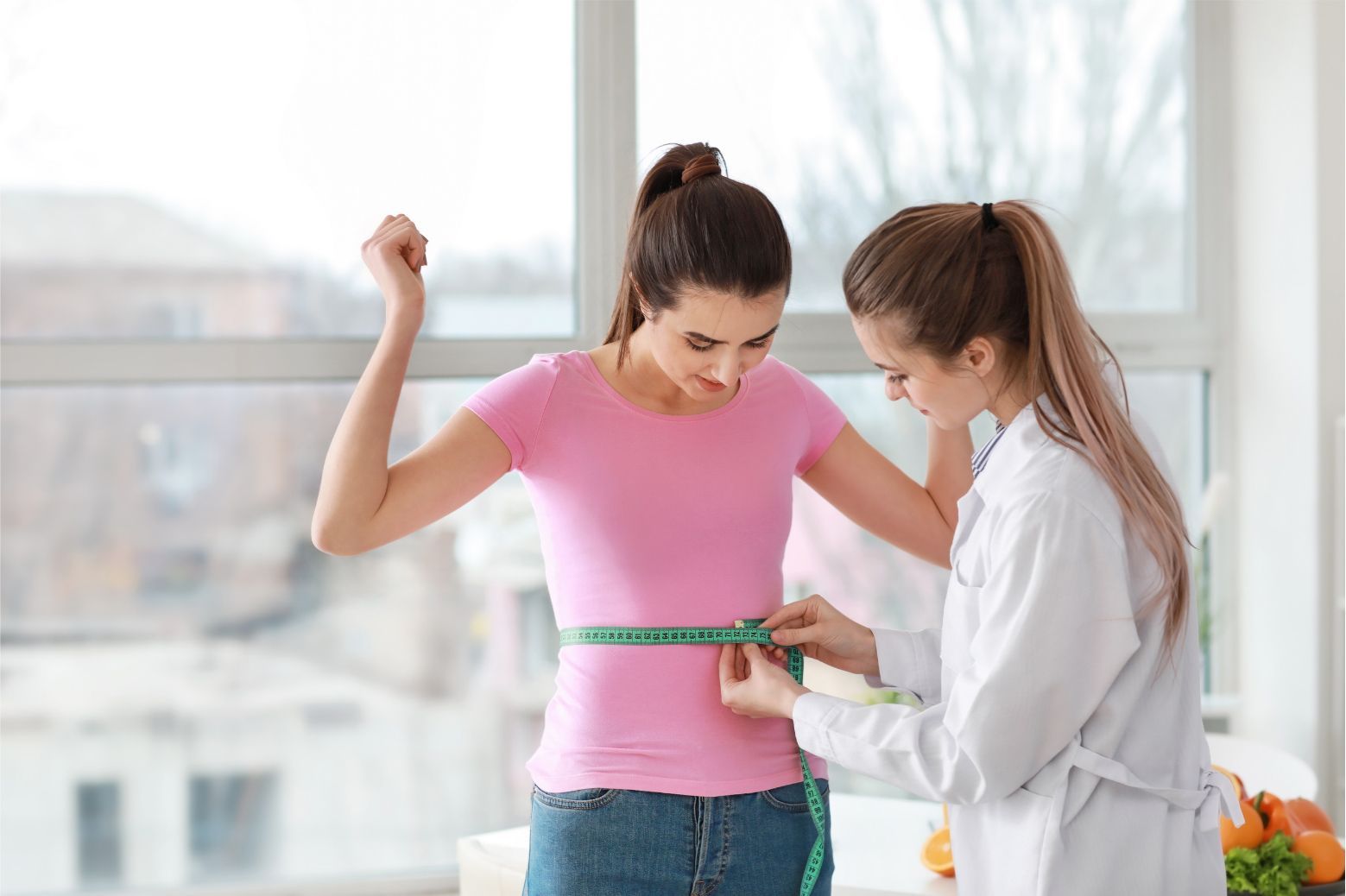 Woman measuring waist while standing in a bright room with a helper holding a tape measure