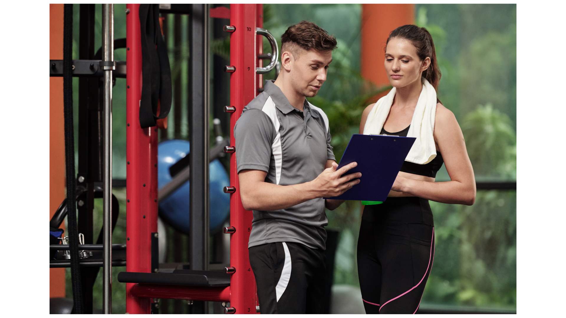 Gym trainer reviewing workout plan with a client beside exercise equipment.