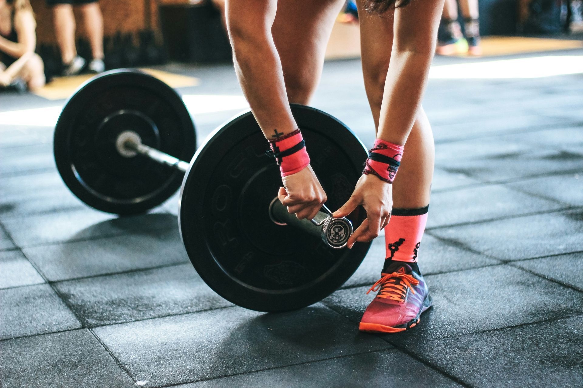 Person lifting a barbell in a gym, wearing black shorts, red wraps, and bright orange shoes.