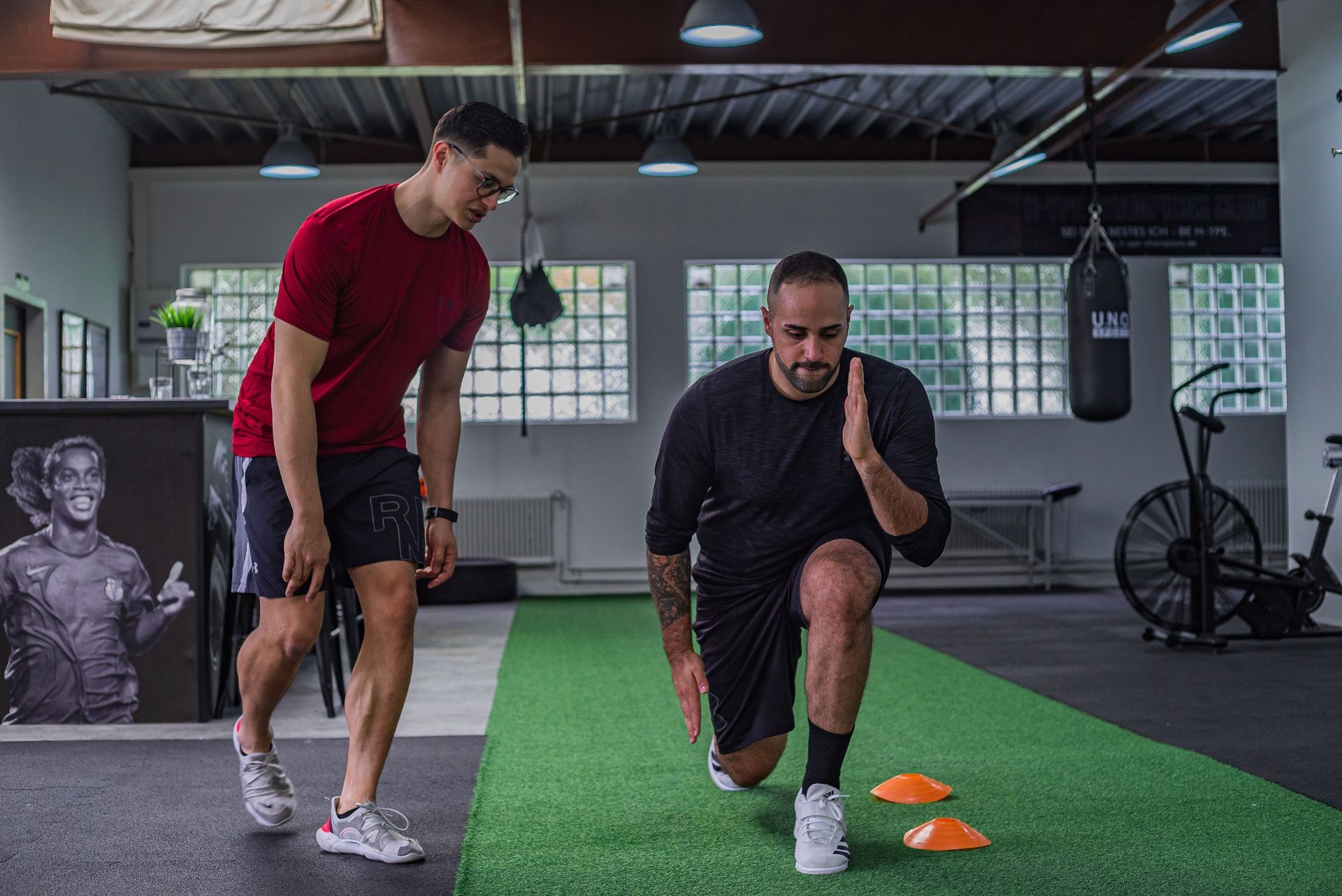 Two people doing lunges in a gym beside orange cones and a punching bag
