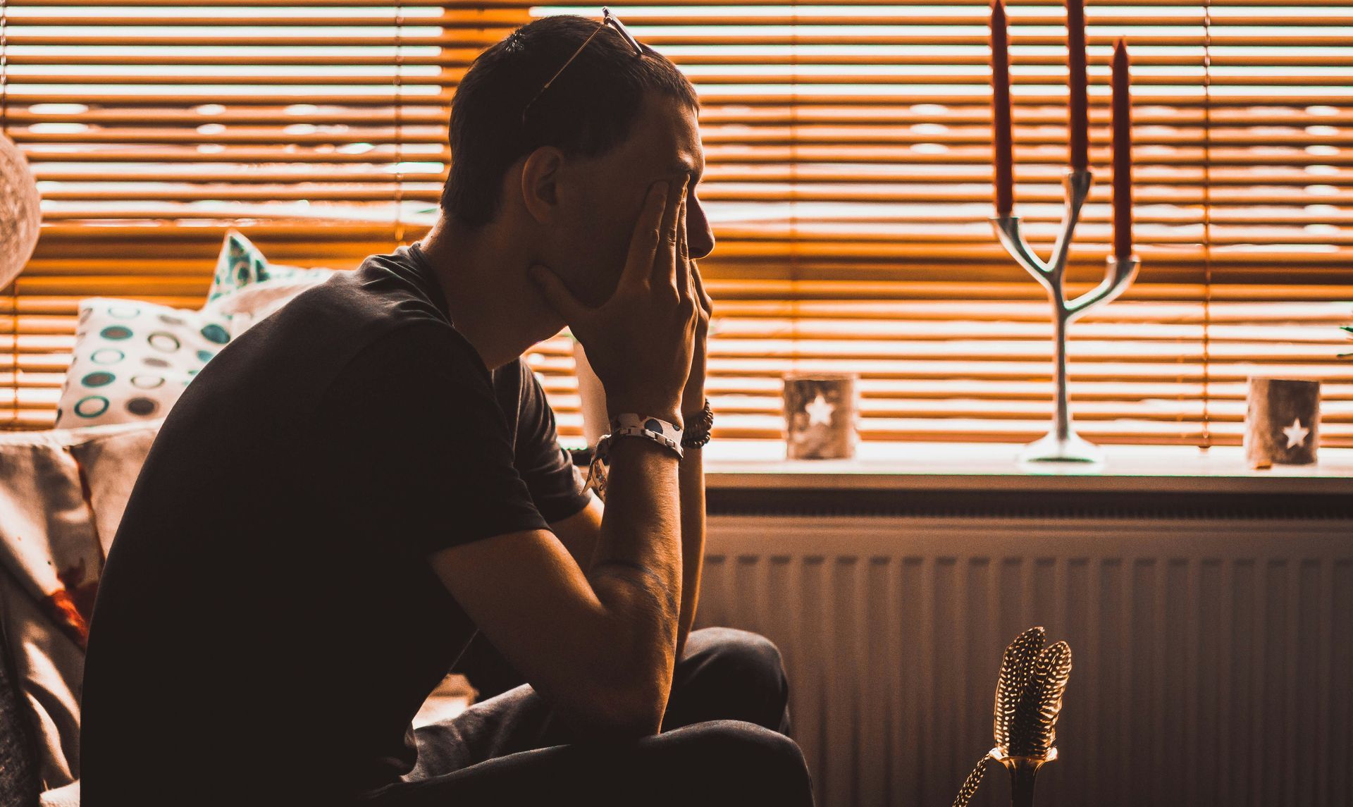 Silhouette of a person sitting by a window, resting their head in their hand, with warm blinds and a guitar nearby