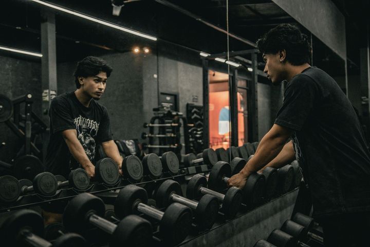 Two men lifting dumbbells in a dim gym, facing each other by a mirrored weight rack.