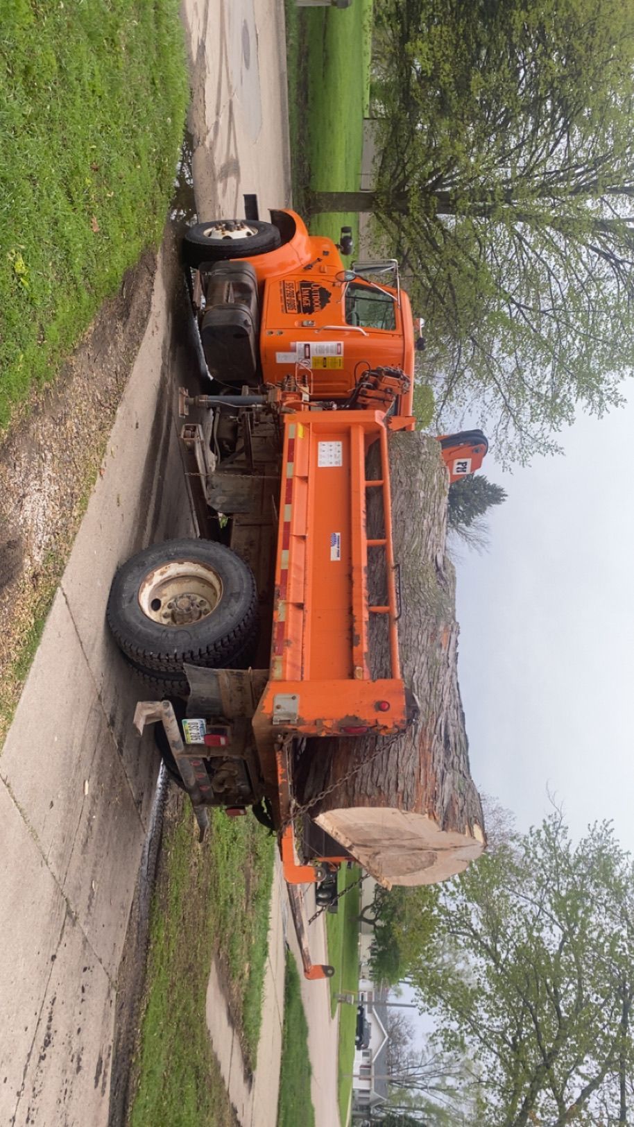 Orange utility truck with a front-mounted digger parked beside a muddy roadside and trees