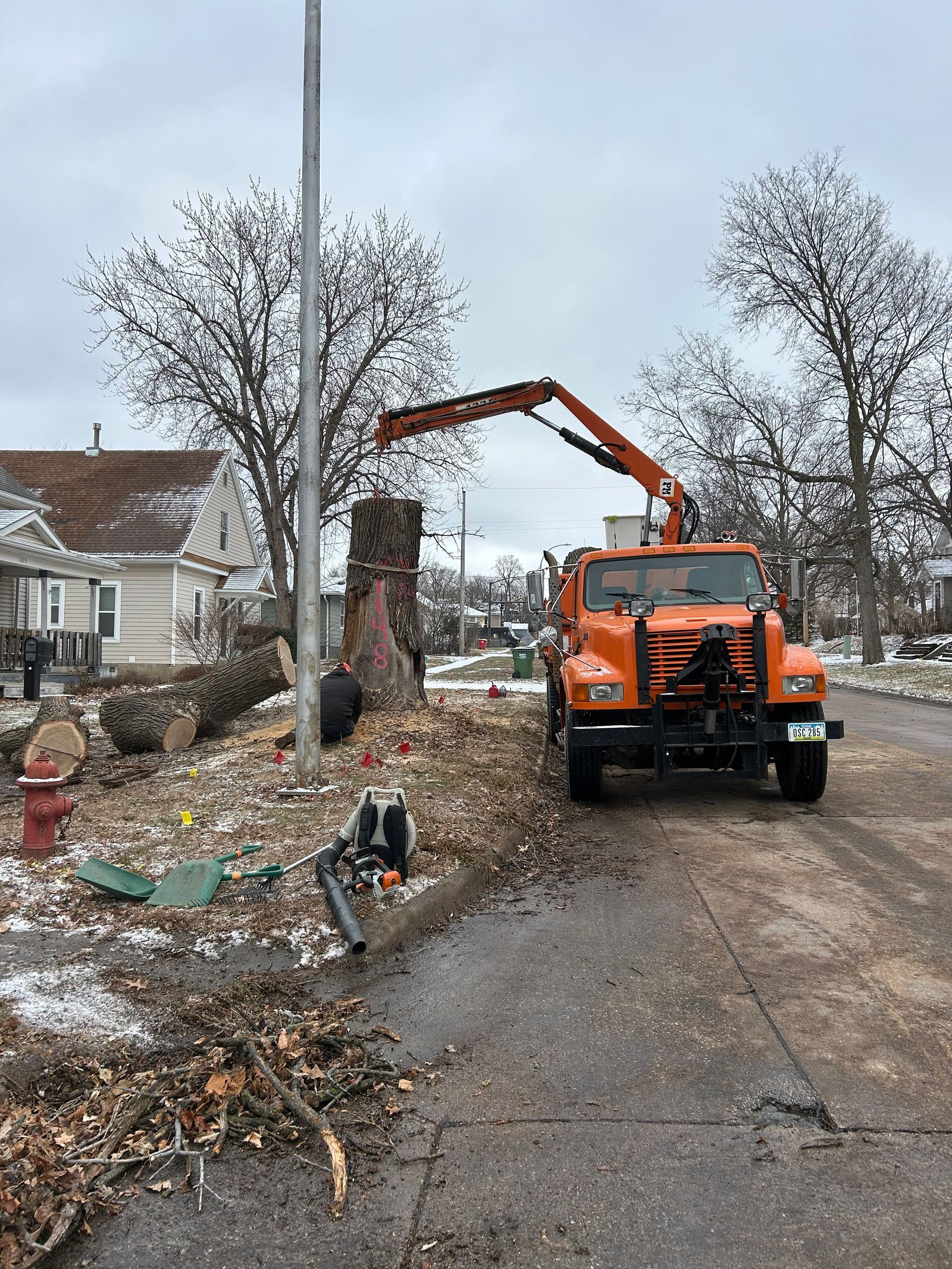 Orange excavator clearing debris beside a house on a damaged street in winter.